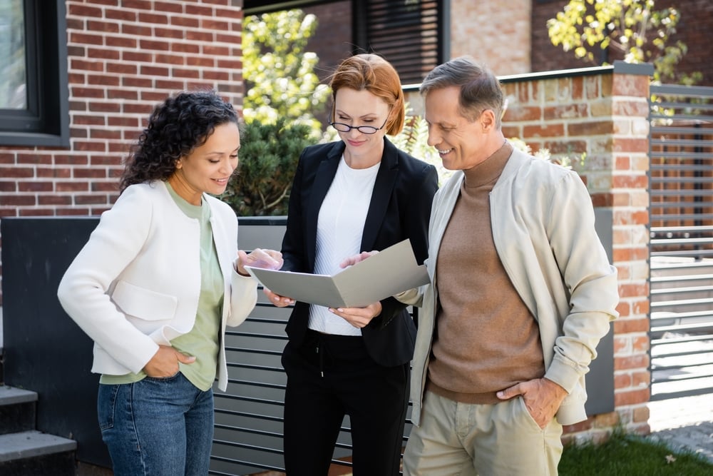 A professional real estate agent showing a folder of documents to a couple while standing outside a modern brick house.