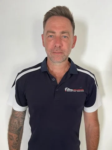 A man with short brown hair and a tattooed arm wearing a navy blue polo shirt with a company logo for a pest and building inspection service.