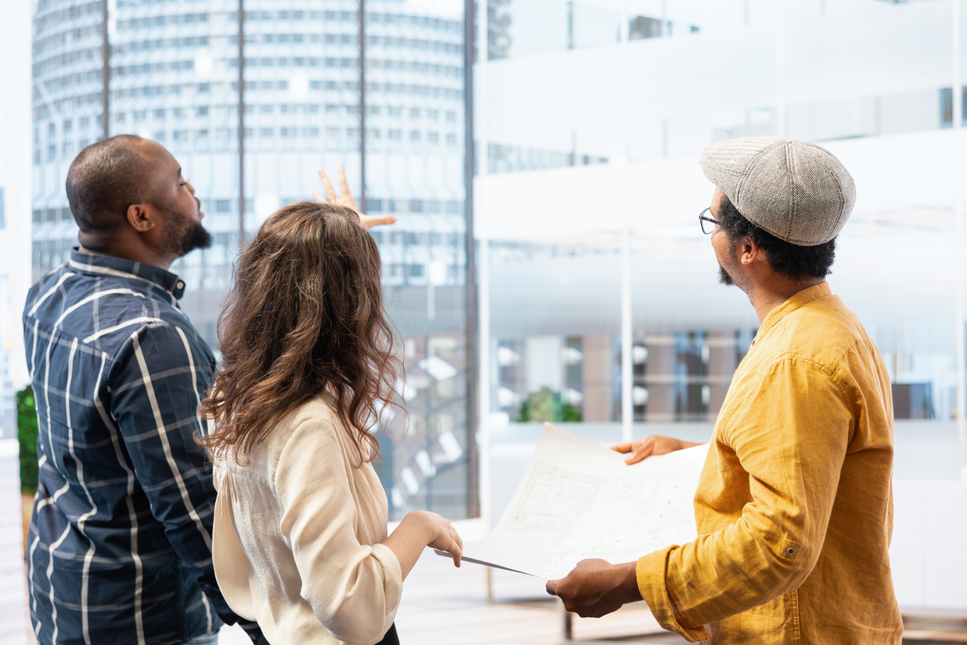 Three people from the back look out a window at a city building while one holds an architectural plan.