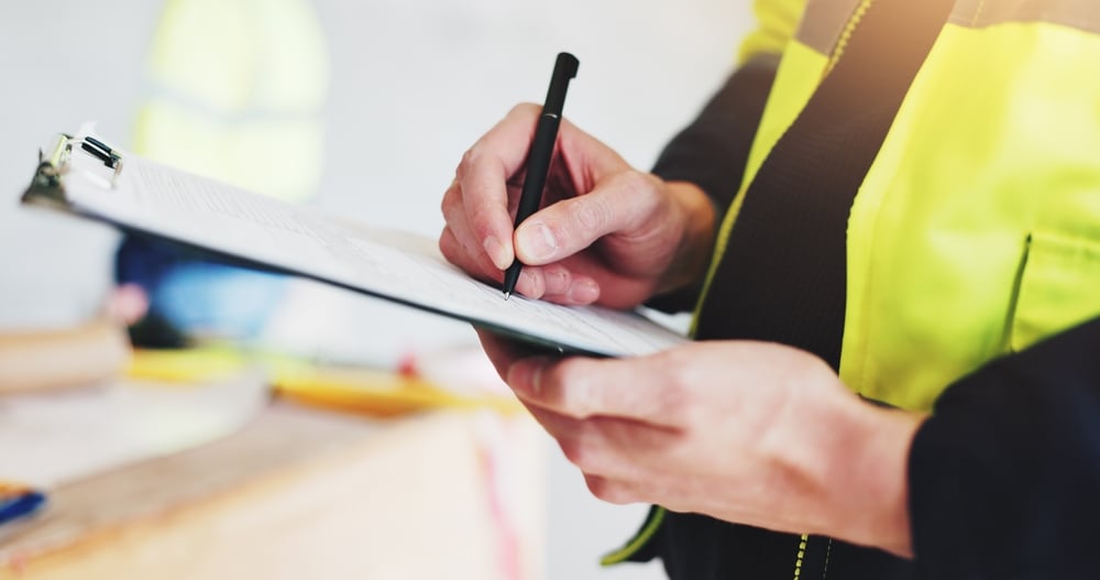 A close-up of a worker in a high-visibility safety vest writing on a document held by a clipboard