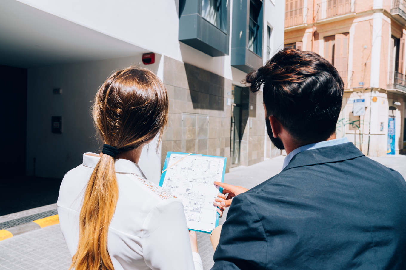A man and a woman viewed from behind, looking at a building plan on a clipboard while standing in front of a modern urban building.