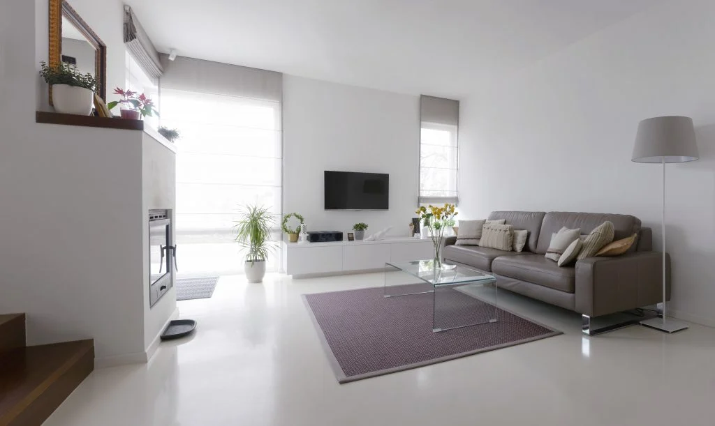 A bright, minimalist living room featuring white walls and floors, a grey leather sofa, a glass coffee table over a purple rug, and a modern fireplace.