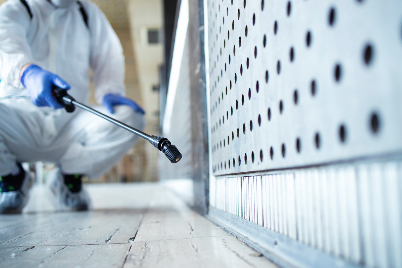 A person in a white hazmat suit and blue gloves uses a spray wand to treat a floor and wall surface
